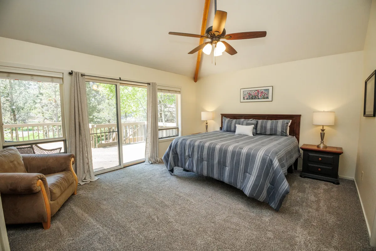 A serene and spacious primary suite featuring a king-size bed with striped bedding, large windows allowing natural light to flood in, a comfortable armchair for quiet moments, and wooden bedside tables. The room is styled for comfort and tranquility.