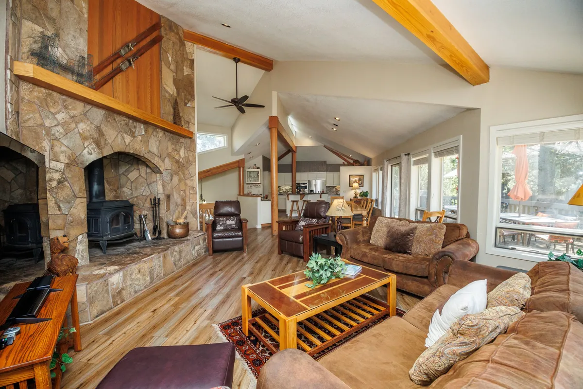 The image showcases a bright, inviting living room with a striking stone fireplace, exposed wooden beams, plush sofas arranged for conversation or relaxation, and expansive windows that look out onto natural scenery. The wooden coffee table and warm lighting create a cozy, rustic-chic atmosphere ideal for gathering with friends or family.