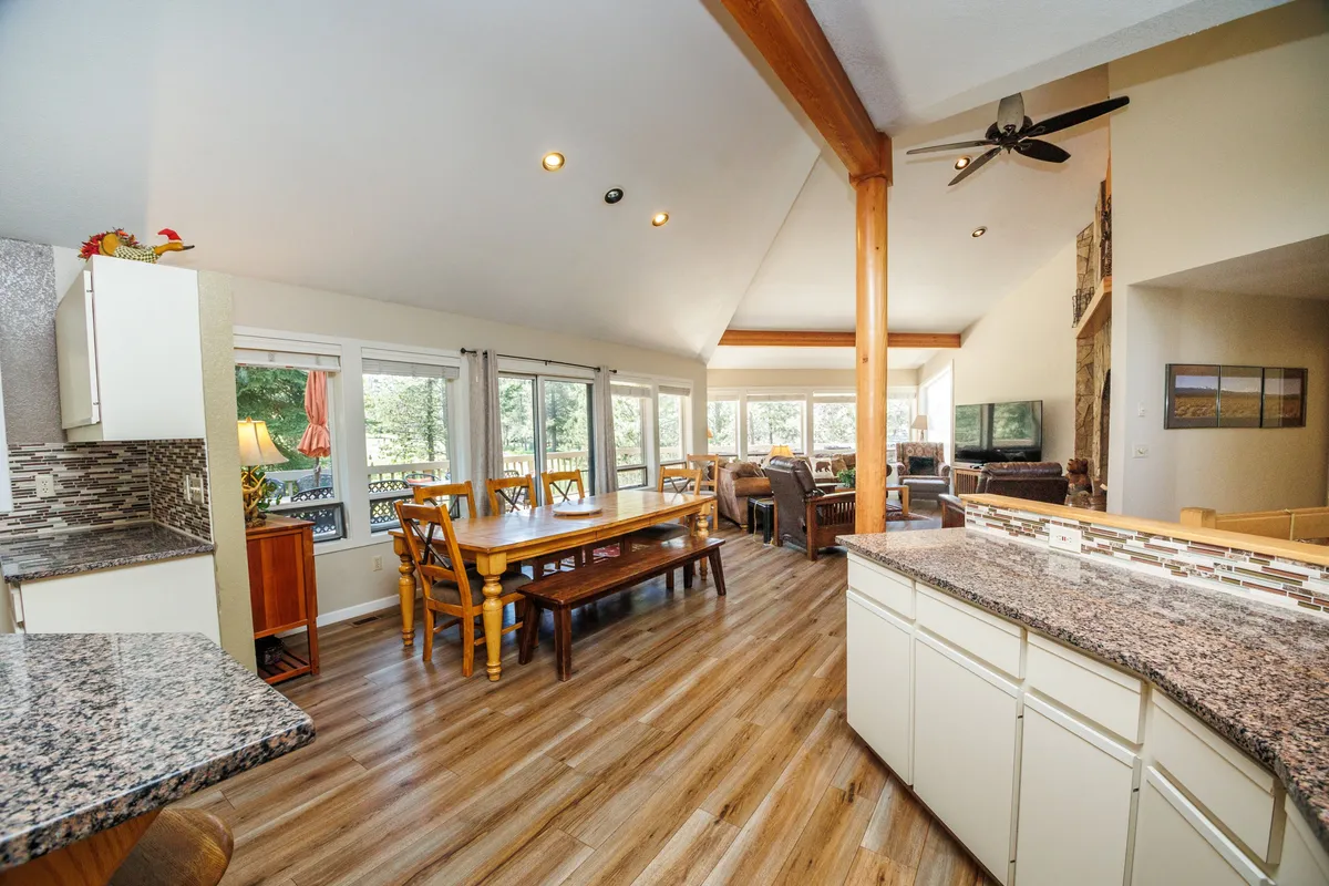 A broad view encompassing the dining area, large windows, living space, and kitchen island seating. Sunlight streams in, highlighting the open-concept design that seamlessly connects lounging, dining, and kitchen spaces.