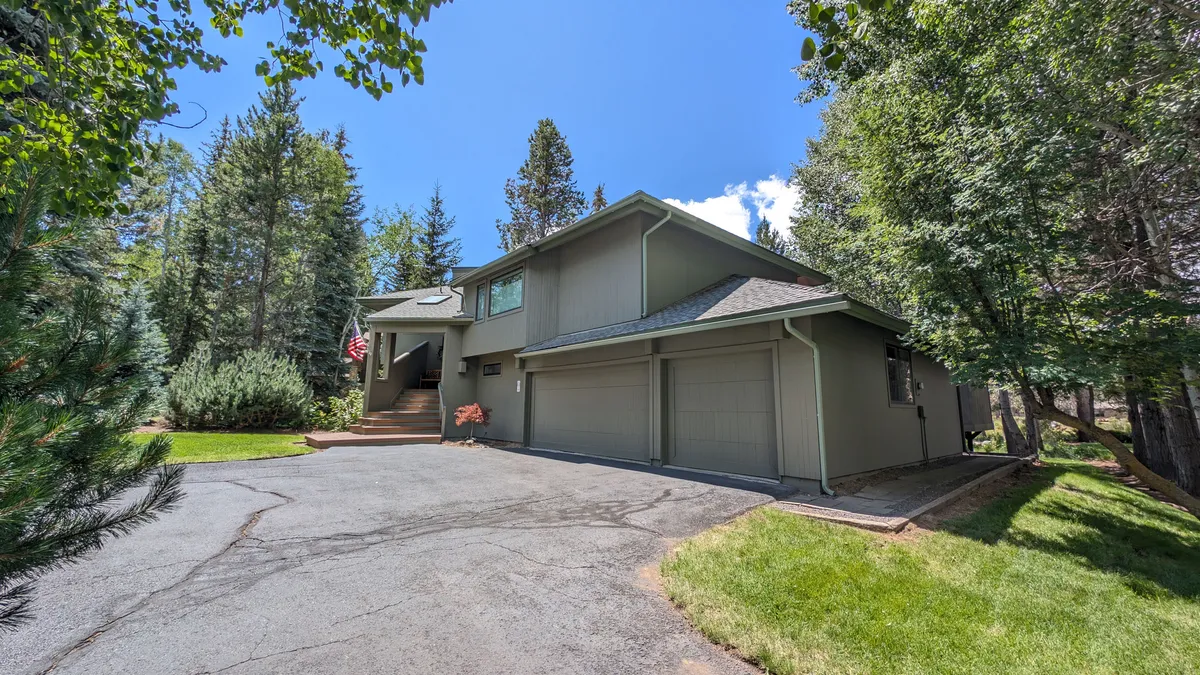 The house’s facade is framed by mature trees, with a welcoming driveway, front steps, and an American flag. The image captures the home’s curb appeal, the sense of arrival, and the serene setting of Sunriver.