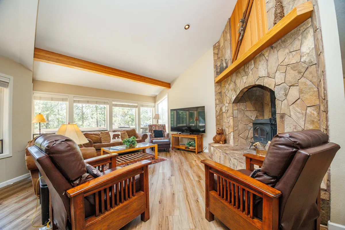 This image showcases a welcoming living area with a prominent stone fireplace, large windows that flood the room with natural light, comfortable seating, and a television. The cozy ambience and open layout make it clear this is a central gathering spot for relaxation and socializing.