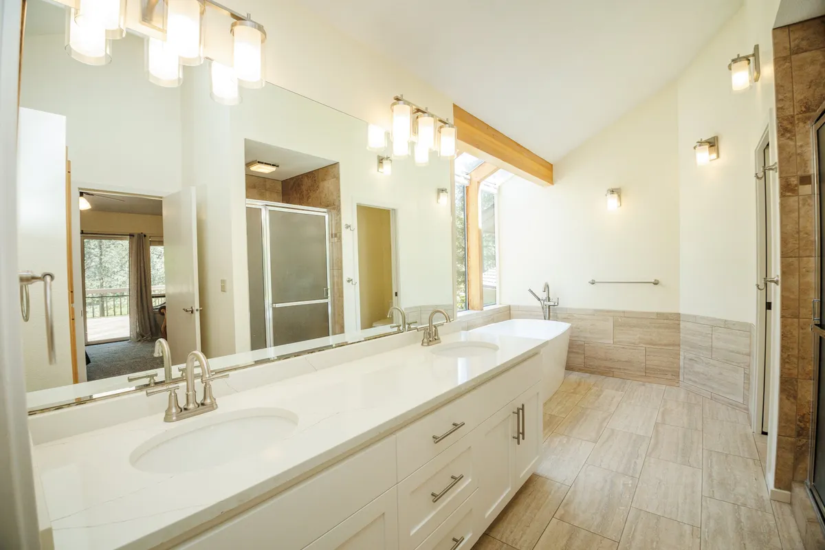 A bright and serene bathroom featuring a double-sink vanity, a spacious bathtub beneath a window, a large mirror, and a walk-in shower with glass doors. The space feels open and calm, ideal for group convenience and relaxation.