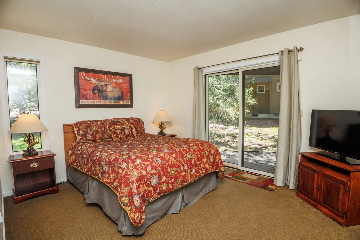 A warm, inviting bedroom featuring a comfortable bed with decorative bedding, framed moose artwork above the headboard, soft lighting from table lamps, and sliding glass doors that look out onto natural greenery. The space is ideal for unwinding and enjoying peaceful views.