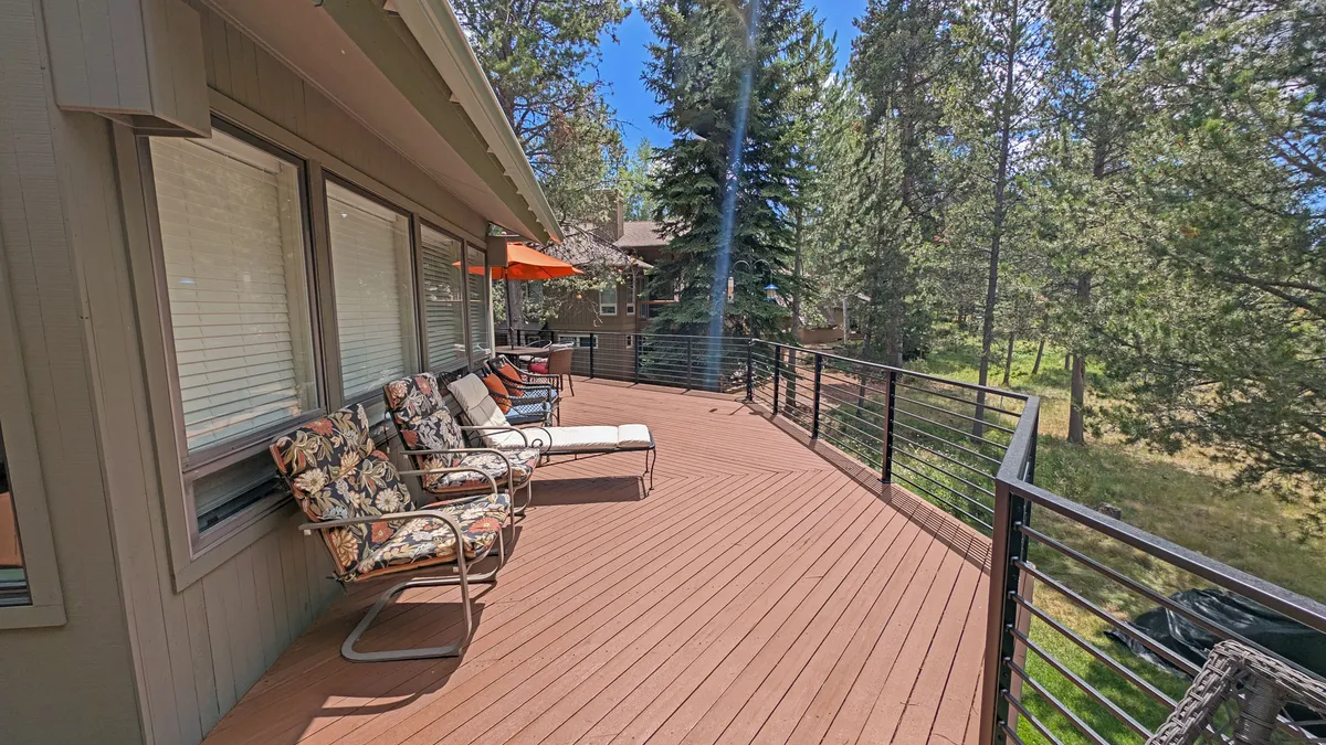 This image shows a spacious deck furnished with comfortable outdoor furniture, surrounded by tall trees and framed by a deck railing with an umbrella for shade. The deck offers a serene spot to relax, dine, or watch the landscape beyond, hinting at the proximity to nature and, potentially, the adjacent golf course.