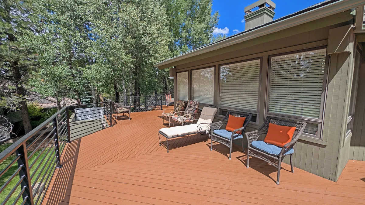 Depicts a cozy outdoor deck seating area framed by mature trees and large windows of the house. The space looks ideal for relaxing, entertaining, or enjoying a beverage while watching the day begin or end. The natural, wooded environment helps set the scene of a getaway immersed in the outdoors.